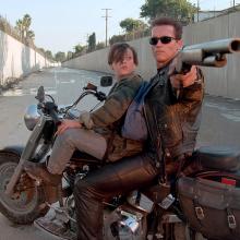 Two individuals sit on a motorcycle one points a gun behind the camera looking stoic.