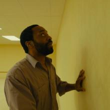 Individual in a yellow, empty room, places his hand on a wall looking at it curiously and afraid.