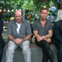 Four men wearing formal-casual attire sit on a ledge looking discouraged, the man on the far left is icing his cheek.