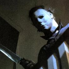 individual in a creepy mask looks down a stairwell with a knife in hand