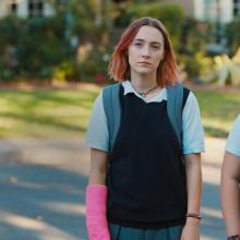 two individuals walk down a street wearing dull school uniforms, looking sad, one has a hot pink cast on