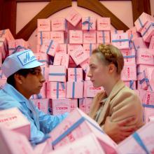 Two individuals sit in a truck full of pink cake boxes looking at eachother