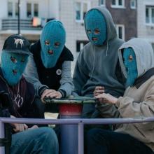 Four young individuals wearing blue ski masks sitting on a playground merry-go-round