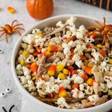 Halloween snack mix with popcorn, candy corn, and pretzels, and spiders and pumpkins on counter in background.