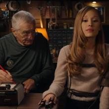 Two individuals sitting down in a dim room. One is strapped to a lie detector machine and the other is examining them and writing on a piece of paper