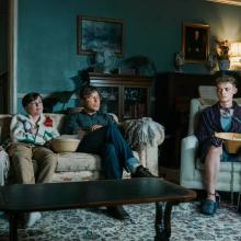 3 indivuals sit on a couch watching tv, one other individual is on a chair tied up, chains around his neck and hands.