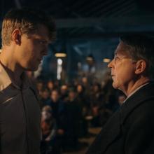 Two people face eachother in a dimly lit room with others watching intently in the background.