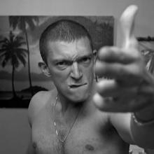 A shirtless man with a buzz cut and a chain necklace glares at the camera while extending his middle finger toward it. The black-and-white image uses a close perspective that makes his hand appear oversized in the foreground.