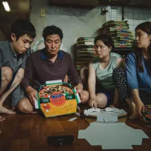 Four Korean family members sit on the floor in a cramped room surrounded by stacks of pizza boxes. The father holds a pizza box while the others look at it with serious, concerned expressions. An unfolded pizza box lies on the floor in front of them.