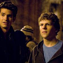 Two young men stand side by side in a dimly lit outdoor setting at night, surrounded by a crowd. One wears a dark coat, while the other wears a blue T-shirt under a brown hoodie. Both look off-camera with serious, attentive expressions, illuminated by warm golden lighting.
