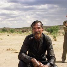 A mustached man in a dark suit crouches in the foreground of a dry, open landscape with a pipe in his mouth. Two men stand behind him on either side, one wearing a cowboy hat and the other with his arms crossed. Sparse vegetation, small buildings, and a cloudy sky stretch across the background.